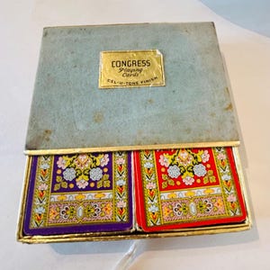 May include: A vintage Congress Playing Cards box with a light blue lid and a gold label reading CONGRESS Playing Cards CEL-U-TONE FINISH. The box contains two decks of cards, one with a purple floral back design and one with a red floral back design.