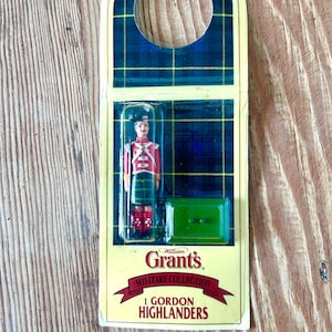 May include: A vintage William Grant's Military Collection figurine of a Scottish Highlander soldier in a red jacket, green kilt, and black hat. The figurine is in a clear plastic case with a green base, set against a plaid background. The packaging includes the text "I. Gordon Highlanders."