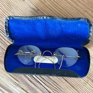 May include: A pair of gold-tone rimless spectacles with round lenses and thin wire temples, resting inside a black hard-shell case lined with vibrant blue velvet fabric. The spectacles feature a delicate bridge and small nose pads.