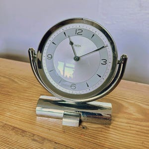 May include: A silver-toned, vintage Smiths desk clock with a round face. The clock has a light gray face with black numerals and hands. The clock is supported by a rectangular base and curved supports. The clock is on a wooden surface.
