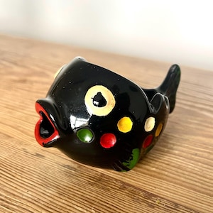 May include: A glossy black ceramic fish-shaped bowl with a wide open mouth painted in bright red. The body features a large pale yellow eye and several small, colorful polka dots in shades of yellow, red, green, and white across its rounded surface.