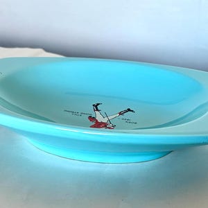 May include: A light blue ceramic serving dish with a shallow, oval shape. The dish features a decorative illustration of a person in a red and white outfit, along with the text "BORN 1820 - STILL HOME STRONG".