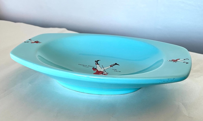 May include: A light blue ceramic serving dish with a shallow, oval shape. The dish features a decorative illustration of a person in a red and white outfit, along with the text "BORN 1820 - STILL HOME STRONG".
