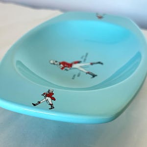 May include: A light blue, oval-shaped dish with a glossy finish. The dish features several small illustrations of a person in a red coat and white pants. The dish is likely vintage and suitable for serving snacks or as a decorative piece.