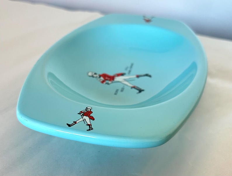 May include: A light blue, oval-shaped dish with a glossy finish. The dish features several small illustrations of a person in a red coat and white pants. The dish is likely vintage and suitable for serving snacks or as a decorative piece.