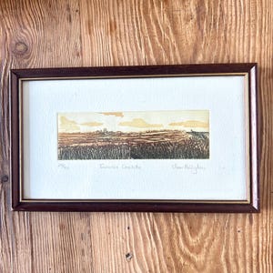 May include: Framed landscape art print with a brown wooden frame. The print depicts a field with a town in the distance under a yellow sky. The text "Towards Cornhill" is visible, along with the artist's signature and the number 29/60.