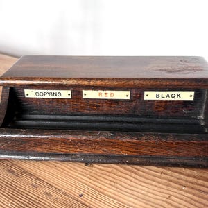 May include: A vintage wooden desk organizer with three labeled sections: "COPYING", "RED", and "BLACK". The organizer is made of dark wood with a rectangular shape and a small tray at the bottom. The labels are on small rectangular plates.