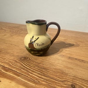 May include: A small, ceramic Poole pottery pitcher with a rounded body and a handle. The pitcher is primarily cream-colored with a dark brown rim and handle. The design features a brown and green landscape with the word "Poole" written on the front.