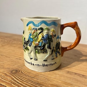 May include: A cream-colored ceramic pitcher featuring a raised, colorful relief of figures riding a horse. The handle is brown, and the text "Widecombe-in-the-Moor" is printed in black along the base. A blue wavy line decorates the top rim.
