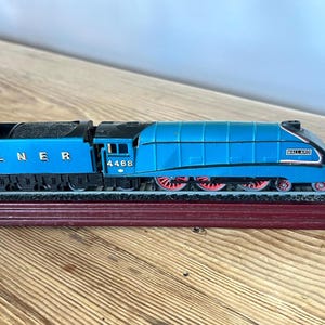 May include: A blue LNER 4468 Mallard model steam locomotive train with red wheels, black detailing, and a coal tender. The train is mounted on a section of model railway track attached to a dark brown rectangular wooden display base.