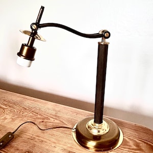 May include: A desk lamp featuring a round brass base, a vertical ribbed black column, and an adjustable curved black arm. The lamp includes a brass socket housing, a white bulb fitting, and a brown power cord with an inline switch attached.