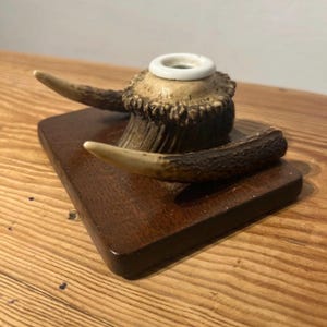 May include: A desk accessory featuring a dark brown rectangular wooden base topped with a natural textured antler piece. A smooth white ceramic ring is embedded in the centre of the antler, creating a functional holder for a pen or small item.