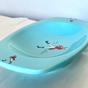 May include: A light blue ceramic dish with a slightly curved rectangular shape. The dish features illustrations of a person in a red and white uniform, with text that reads "Still Going Strong".