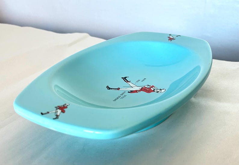 May include: A light blue ceramic dish with a slightly curved rectangular shape. The dish features illustrations of a person in a red and white uniform, with text that reads "Still Going Strong".