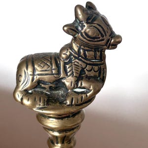 May include: A brass figurine of a reclining bull with etched geometric patterns on its body, mounted on a circular pedestal base. The metal has a polished finish with dark, recessed areas highlighting the engraved details on the animal.