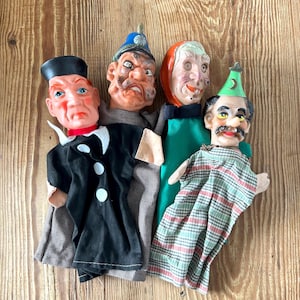 May include: Four vintage hand puppets with painted faces and fabric clothing. The puppets include a man in a black suit, a stern-faced man in a blue hat, a woman with a red head covering, and a man in a plaid robe and green hat.