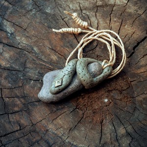 May include: A pendant featuring a sculpted, sage-green clay snake wrapped around a smooth, dark gray river stone. The snake has small indentations along its body and a diamond-shaped metallic accent. It hangs from a light tan, knotted twine cord.