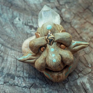Könnte beinhalten: Eine skulptierte braune Kreaturenfigur mit strukturierter Oberfläche, die einen klaren, spitzen Kristall an der Oberseite, kleine goldene Metallkugeln als Augen und hellblaue Farbakzente auf Stirn, Nase und Ohren sowie ein kleines Muscheldetail aufweist.