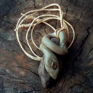 May include: A pendant featuring a sculpted light brown snake wrapped around a dark brown stone. The snake has a small square brass-colored inlay on its head. The piece is suspended from a light-colored, thin, twisted fiber cord necklace.
