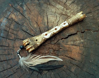 May include: A carved, light-colored cylindrical object with a textured surface and multiple small holes along its length. One end features a decorative attachment with a small black bead, a silver-toned metal component, and two feathers.