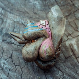 Könnte beinhalten: Ein skulptierter Kreaturenkopf mit brauner, texturierter Oberfläche, rosa Akzenten, blauen Streifen und einem einzelnen orangefarbenen Auge. Ein großer, klarer, spitzer Quarzkristall ist an der Seite befestigt, und ein kleiner quadratischer Metallniet ist in den rosa Bereich eingelassen.