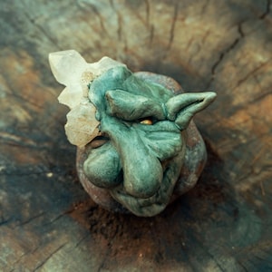 Könnte beinhalten: Eine grüne Trollskulptur mit einem Kristall am Kopf. Der Troll hat eine Knollennase, spitze Ohren und einen strengen Gesichtsausdruck. Die Skulptur befindet sich vor einem strukturierten Holzgrund.