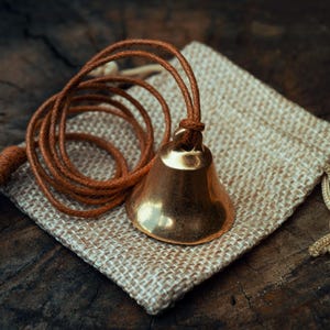 May include: A small, polished brass bell with a brown leather cord, resting on a hessian pouch. The bell has a rounded shape and a warm, golden hue. The cord is coiled around the bell, and the pouch is beige.