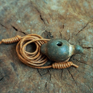 Può includere: Un fischietto in ceramica verde scuro con un foro nella parte superiore, attaccato a un cordino marrone. Il fischietto è appoggiato su un pezzo di legno.