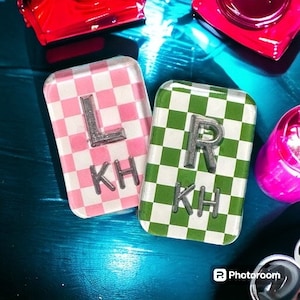 Op de afbeelding: Twee vierkante tegels met een geruit patroon. Eén tegel is roze en wit met de letter "L" en de letters "KH" erop. De andere tegel is groen en wit met de letter "R" en de letters "KH" erop.