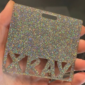 May include: A silver glitter card with the word "XRAY" cut out in the center. The card has a hole at the top for hanging.