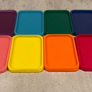 Puede incluir: Ocho bandejas rectangulares en varios colores, incluyendo rosa, amarillo, naranja, rojo, azul, verde y morado. Las bandejas están dispuestas en una fila sobre una superficie gris claro.