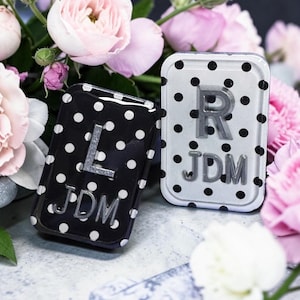 May include: Two rectangular, polka-dotted containers with silver lettering. One is black with white dots and the letter "L" and the initials "JDM". The other is silver with black dots, the letter "R", and the initials "JDM". Pink flowers are in the background.