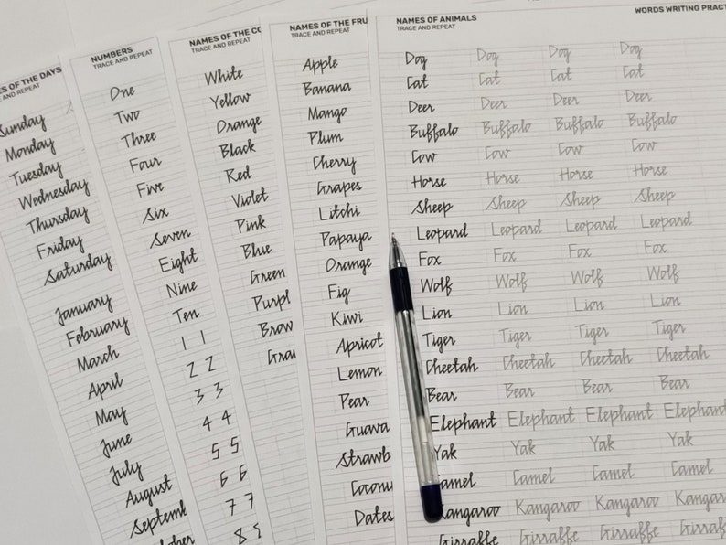 May include: A collection of handwriting practice sheets with various topics, including days of the week, numbers, colors, fruits, and animals. A black and silver pen rests on the pages. The sheets are designed for tracing and repeating words.