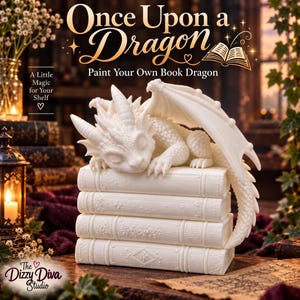 May include: A white, unpainted dragon figurine resting on a stack of four books. The text "Once Upon a Dragon" and "Paint Your Own Book Dragon" appears above the dragon. The text "A Little Magic for Your Shelf" and "The Dizzy Diva Studio" is visible.