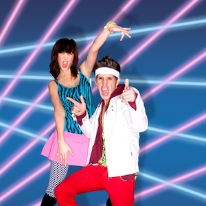 May include: A man and a woman dressed in 1980s style clothing pose in front of a blue background with pink and blue laser lights. The woman is wearing a zebra print tank top, a pink skirt, and black leggings. The man is wearing a white jacket, red pants, and a headband.