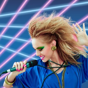 May include: A woman with large, curly blonde hair sings into a microphone. She is wearing a blue dress with a gold necklace and a gold earring. The background is a blue and pink laser light show.
