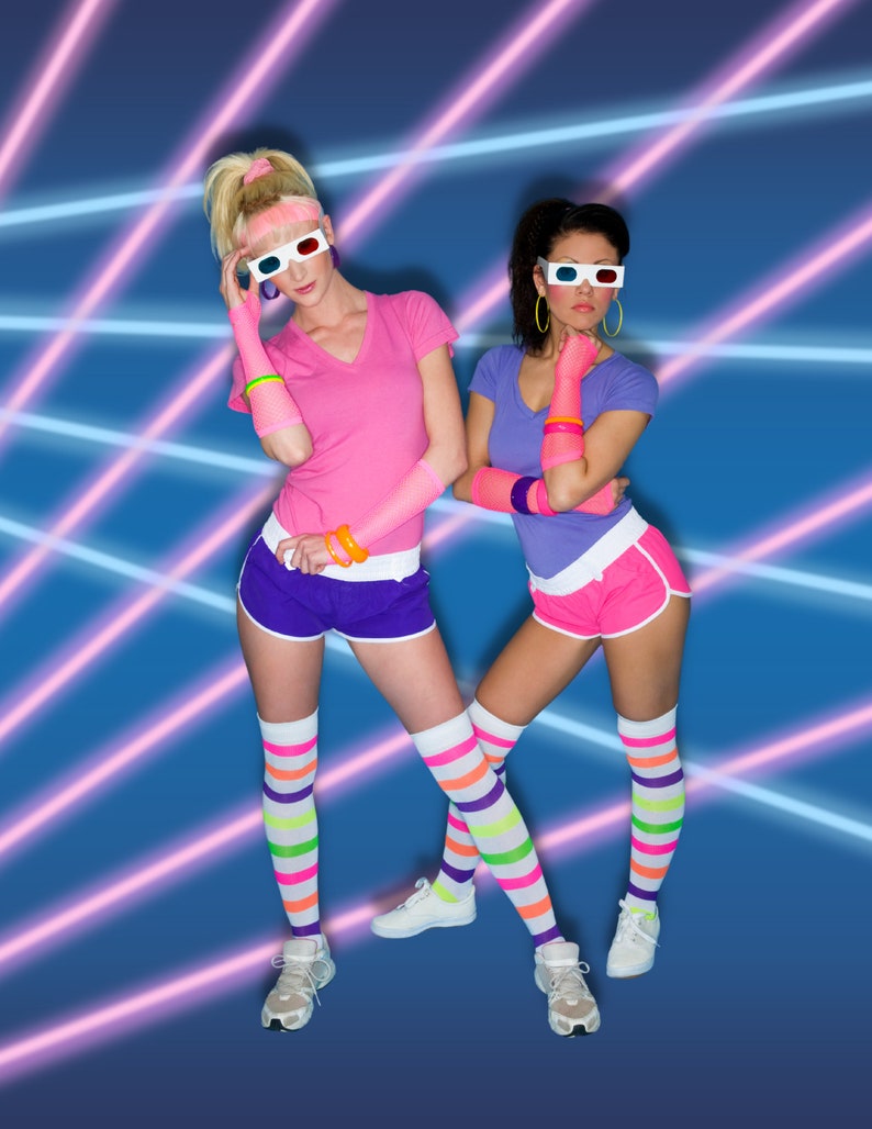 May include: Two women wearing colorful 80s style clothing and 3D glasses pose in front of a blue and pink laser background. They are wearing bright pink and purple shorts, striped socks, and white sneakers.