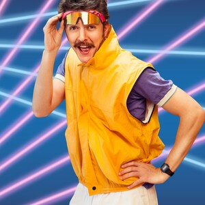 May include: A man wearing a yellow vest, white pants, and a pink and blue headband with sunglasses. He is standing in front of a blue and pink background with neon lights.