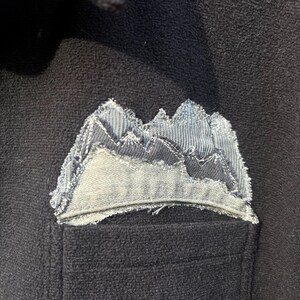 May include: Black button-up shirt with a mountain-shaped patch on the pocket. The patch is made of layered denim in shades of gray and blue, with a distressed, frayed edge. The shirt has black buttons.