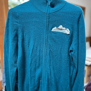 Może przedstawiać: Turkusowy sweter zapinany na zamek błyskawiczny z małą naszywką w kształcie góry na lewej piersi.