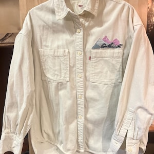 Puede incluir: Camisa de pana color crema con mangas largas, dos bolsillos en el pecho y cierre de botones. La camisa presenta un diseño de montaña bordado en un bolsillo y una pequeña etiqueta roja. La camisa está colgada de una percha negra.