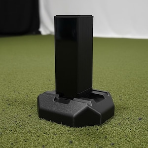 Puede incluir: Un objeto negro, rectangular y vertical con una superficie acanalada, montado sobre una base negra y geométrica. El objeto se encuentra sobre un fondo verde texturizado, lo que sugiere un entorno al aire libre o deportivo.