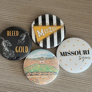 Pode incluir: Quatro botões redondos com temas da Universidade do Missouri. Um é preto com um tigre e o texto BLEED GOLD. Um tem listras pretas e brancas com uma flâmula dourada MIZZOU. Um é branco com pontos dourados e o texto MISSOURI tigers. Um mostra um estádio.