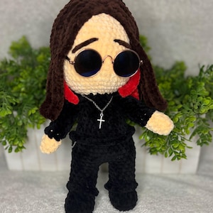 May include: A handmade crochet doll with brown hair, round sunglasses, and a black outfit. The doll has a silver cross necklace and red accents in the hair. The doll is standing in front of a white planter with green foliage.