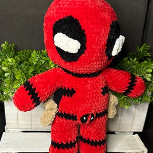 May include: Red and black crocheted plush toy of Deadpool, a superhero character. The toy has a white eye mask and a black belt with a red Deadpool logo.