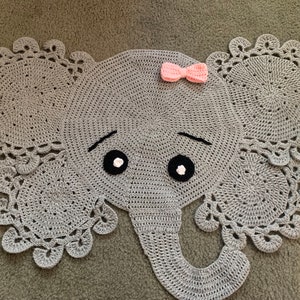 May include: A gray crocheted elephant rug with a pink bow on top. The elephant has black eyes and a gray crocheted trunk.