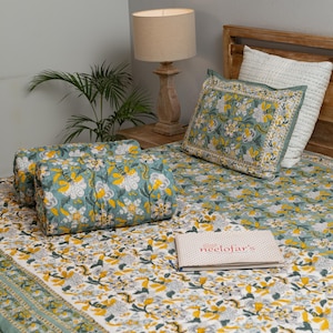 Puede incluir: Una cama con un edredón, almohadas y un cojín a juego con estampado floral. La ropa de cama presenta un diseño floral en verde azulado, amarillo y blanco. Un libro con el texto "neelofar's" está sobre la cama. También se ve una lámpara de madera y una planta.