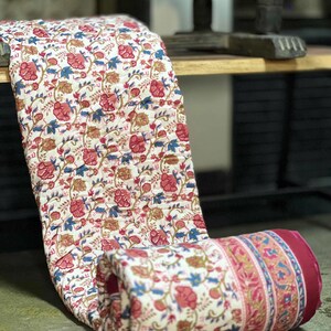 May include: A floral patterned quilt in shades of red, blue, and gold on a cream background. The quilt is rolled up, revealing a burgundy border with a decorative stripe. The quilt is made of a soft, quilted material.