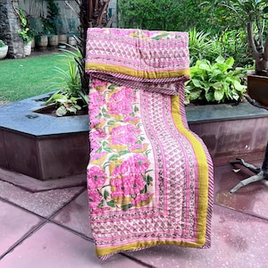 Könnte beinhalten: Eine gesteppte Decke mit einem sich wiederholenden Muster aus rosa Blumen und grünen Blättern auf cremefarbenem Grund. Die Decke hat einen breiten senfgelben Rand und eine schmale gestreifte Kante mit sichtbaren Nähten auf der Oberfläche.