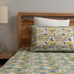 May include: A bed with a wooden headboard and a floral patterned bedspread and pillow. The bedspread and pillow feature a repeating pattern of yellow and white flowers on a light blue background. A white pillow is behind the patterned pillow.